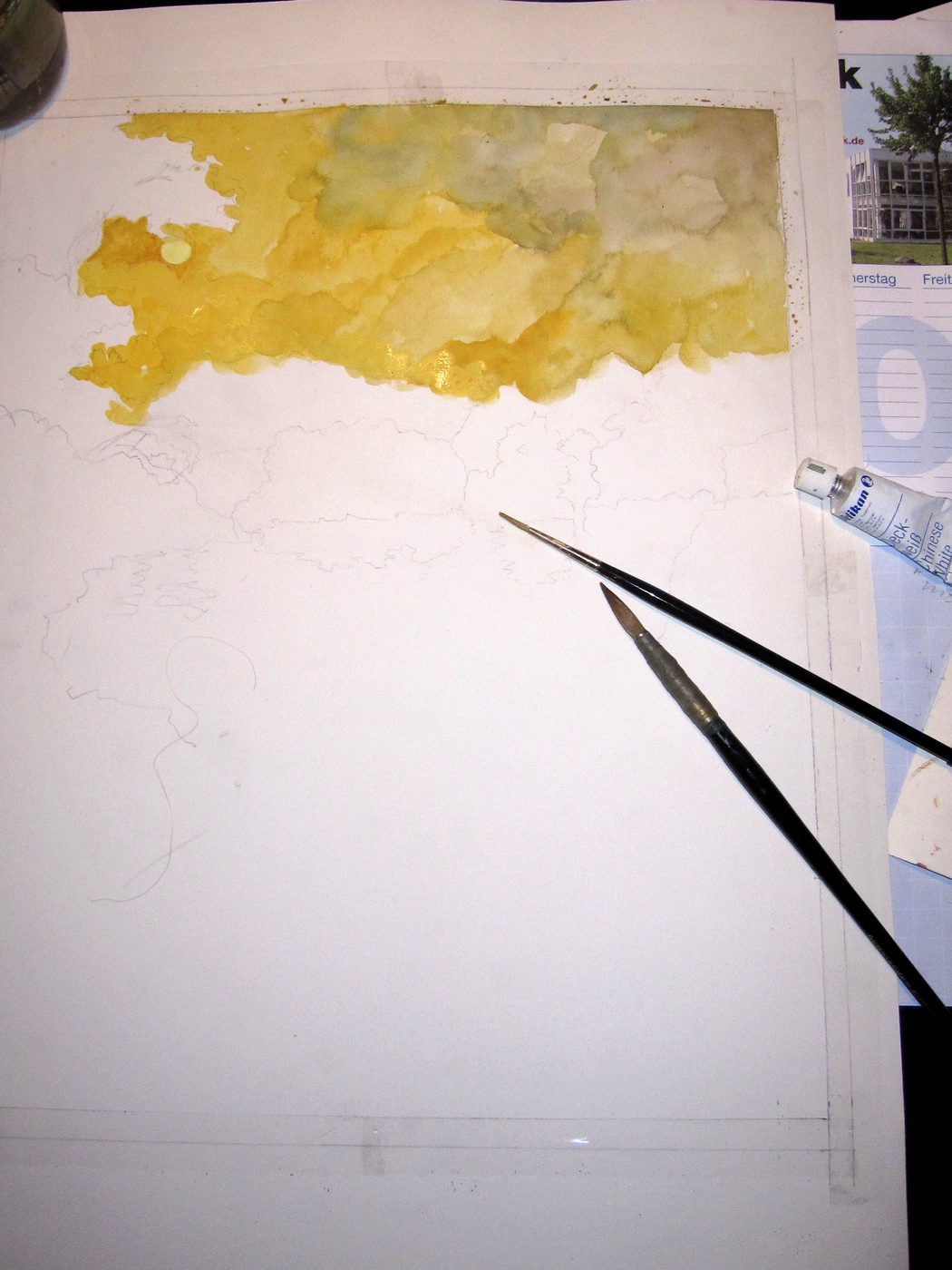 Aquarellpapier auf dem Arbeitstisch, oben eine erste gelbe Himmelsfläche, daneben zwei Pinsel.