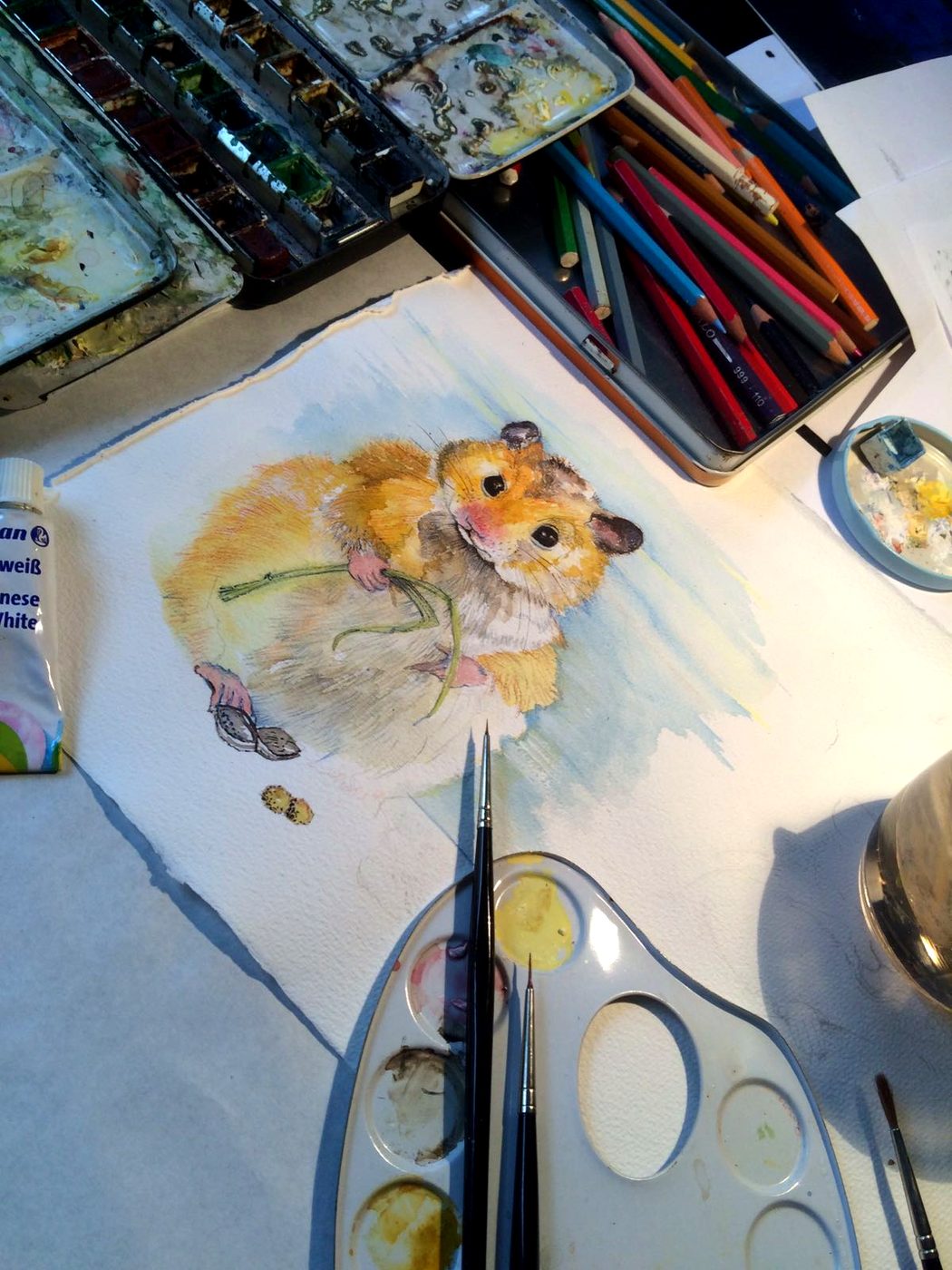 Arbeitstisch mit Hamster-Aquarell, Farbkasten, Buntstiften, Palette und Pinseln.